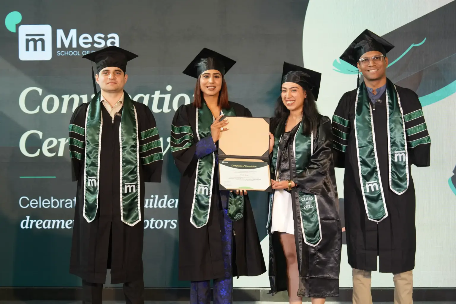 Graduates in gowns on stage at the Mesa School of Business convocation ceremony, one holding a certificate
